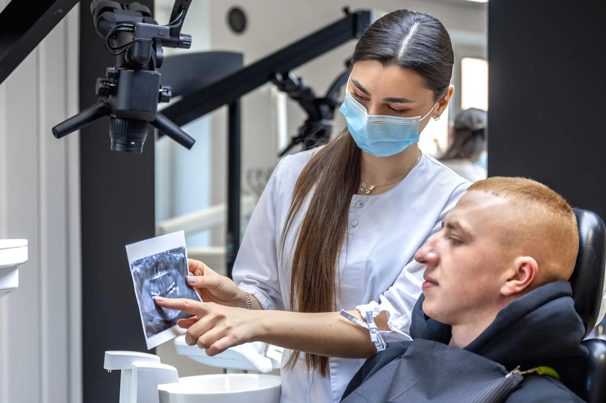 A female dentist wearing a blue surgical mask showing a dental X-ray to a male patient in a modern dental clinic